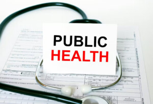 Stock image of a stethoscope on top of a page of gridded paper. The stethoscope is proping up a white card that reads "Public Health" in black and red letters